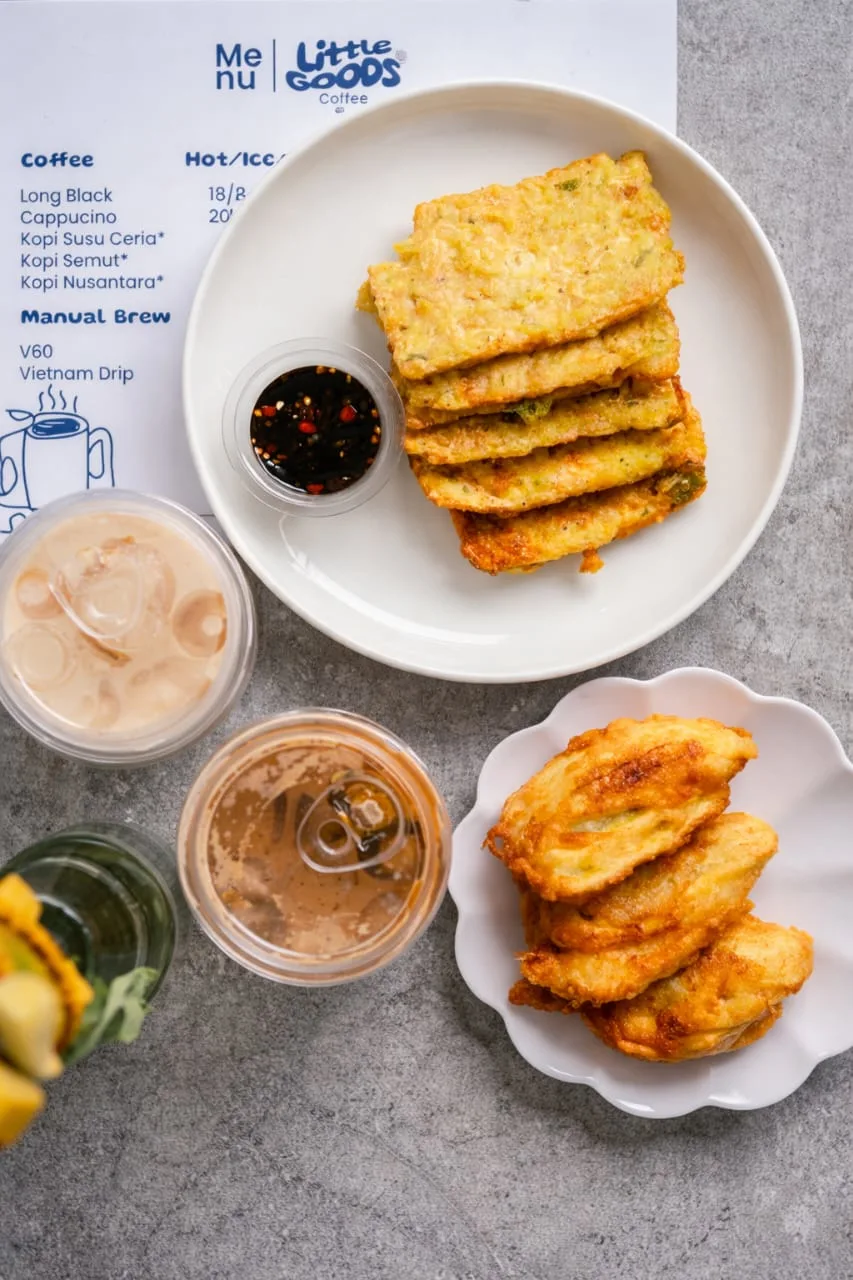 Foto flat lay menu camilan Little Goods Coffee yang terdiri dari sepiring tempe mendoan dengan sambal kecap, sepiring pisang goreng renyah, dua gelas kopi susu dingin, dan lembaran menu di atas meja abu-abu minimalis.