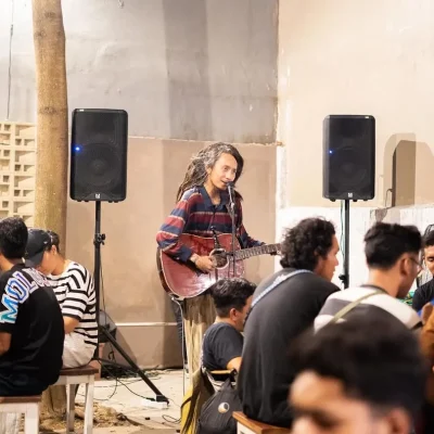 Live Music Session: The Soul of the House 🎸✨ Seorang musisi berambut panjang sedang main gitar akustik dan bernyanyi di depan audiens. Suasana indoor-outdoor yang warm, dengan dua speaker besar di kanan-kiri dan orang-orang yang asyik dengerin sambil nongkrong.
