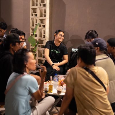 Suasana Hangat Diskusi Komunitas di Malam Hari Sekelompok anak muda sedang duduk melingkar di sebuah meja kecil, berbincang santai dengan ekspresi ceria di sebuah ruangan outdoor atau semi-outdoor dengan latar belakang dinding roster beton.