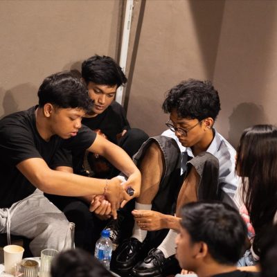 okus Bersama di Tengah Pertemuan Kasual Sekelompok anak muda duduk melingkar di lantai. Seorang pria berbaju hitam fokus mengikat atau memasang sesuatu di pergelangan tangan temannya yang berkacamata, sementara teman lain memperhatikan. Terlihat botol air mineral dan cangkir kertas di latar depan.