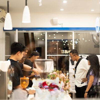 Interaksi Ramah di Balik Meja Kasir Suasana di dalam kafe yang terang, memperlihatkan staf pria berbaju hitam sedang melayani sepasang pelanggan di meja bar. Terlihat buket bunga di latar depan dan lampu gantung minimalis.