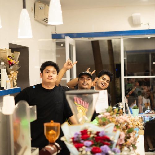 Tiga orang pria berpose ceria di balik meja bar sebuah kafe. Pria di depan memakai kaus hitam polos, pria di tengah memakai kaus Nirvana dan topi, serta pria di kanan tersenyum lebar. Terdapat buket bunga dan peralatan kopi di latar depan.