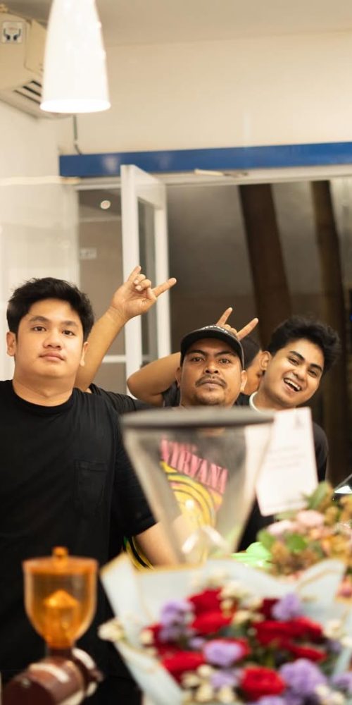 Tiga orang pria berpose ceria di balik meja bar sebuah kafe. Pria di depan memakai kaus hitam polos, pria di tengah memakai kaus Nirvana dan topi, serta pria di kanan tersenyum lebar. Terdapat buket bunga dan peralatan kopi di latar depan.